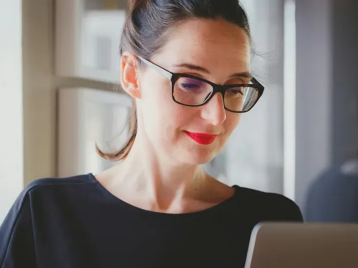 Focused woman in glasses and red lipstick working on a laptop near a window in a bright modern office